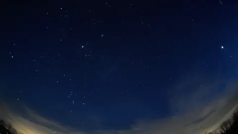 Getty Images A stunning wide-angle nighttime photograph of the evening sky, featuring four planets.
Mars on the left, Jupiter in the middle, and Saturn and Venus on the right. Wispy clouds stretch across the lower part of the sky, with the silhouettes of trees and city lights glowing faintly.