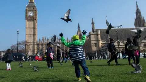 AFP via Getty Images A group of children standing and running around on green space in front of the Houses of Parliament, with one boy in a coat and hat chasing away two pigeons.