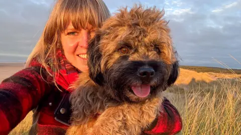 Victoria Gill/BBC News A woman in a red jacket cuddles a small, dog. The dog is looking into the distance and has its tongue out and looks happy. There is a beach in the background and it is a sunny day. 
