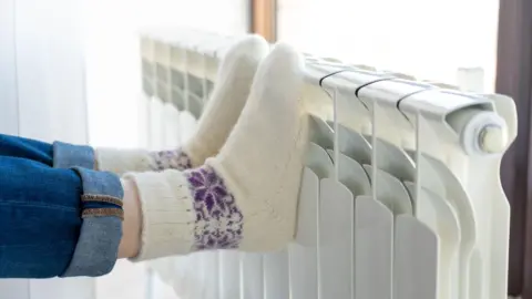 Getty Images A woman wearing rolled-up blue jeans and white and purple knitted socks rests her feet on a white radiator. 